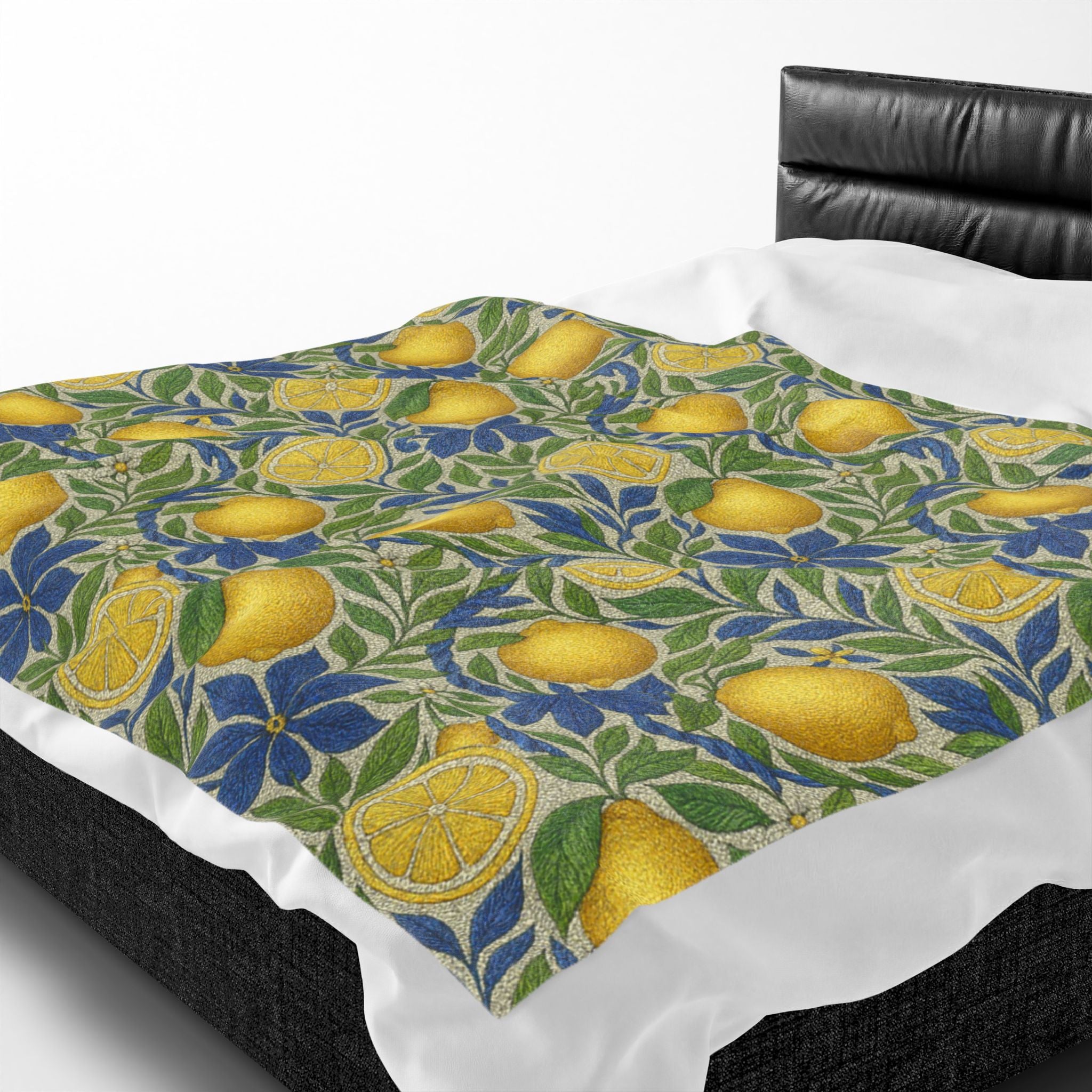 A soft velveteen plush blanket with a yellow lemon and blue flower pattern draped over a bed with white sheets and a black leather headboard.