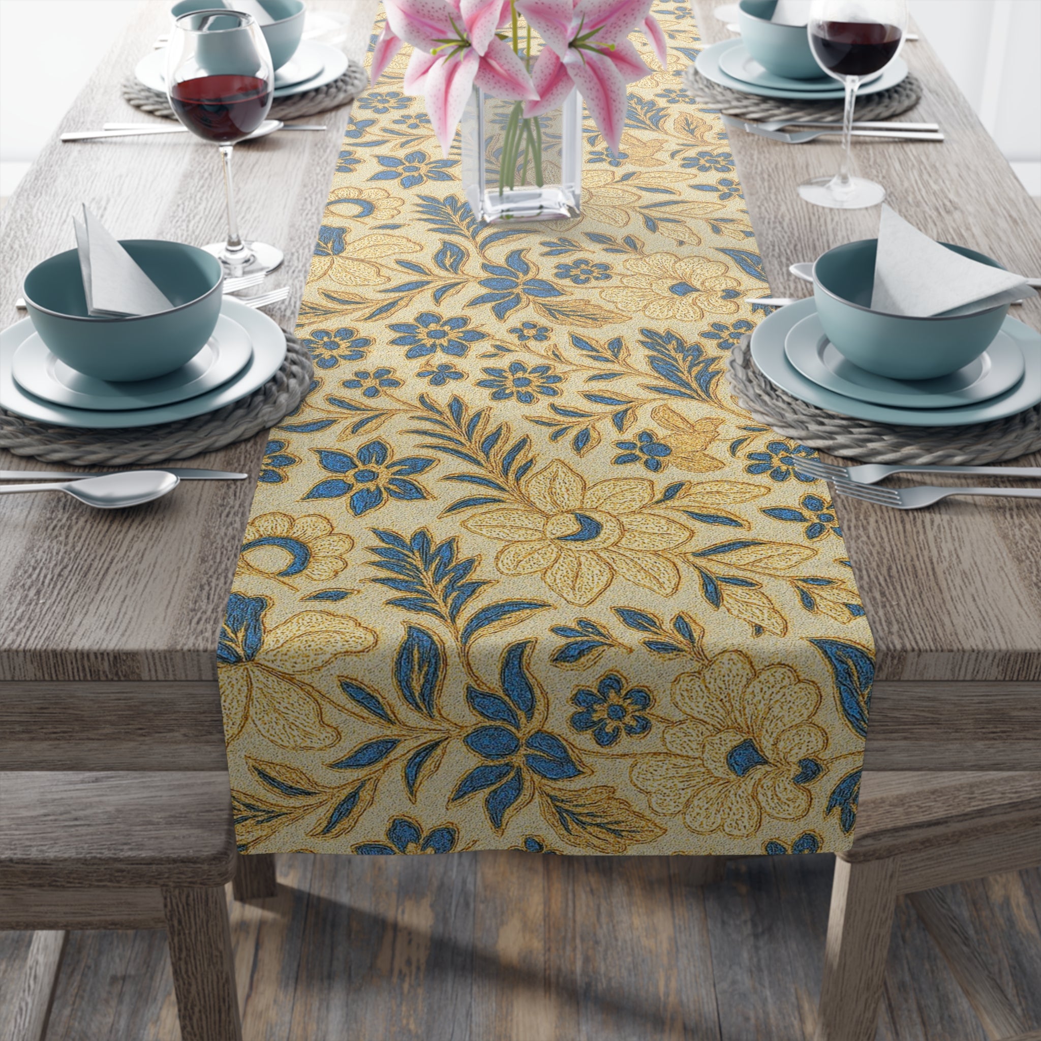 A long table runner with a blue and gold vintage floral pattern inspired by William Morris, displayed on a wooden dining table with blue plates, wine glasses, and pink lilies in a vase.