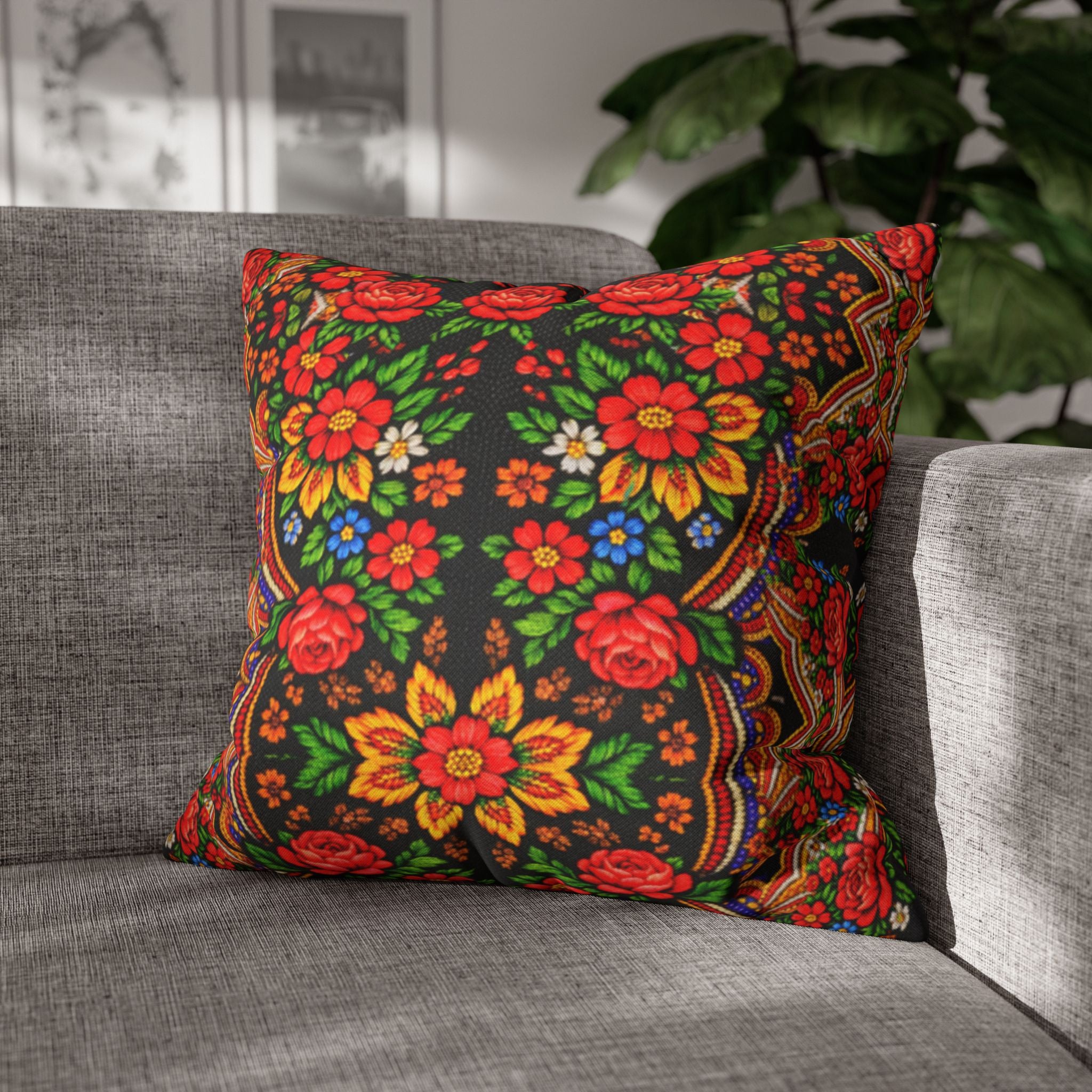 A square decorative pillow named "Mother’s Bloom" with a vibrant red and yellow Ukrainian folk floral pattern on a black background, placed on a grey modern sofa.