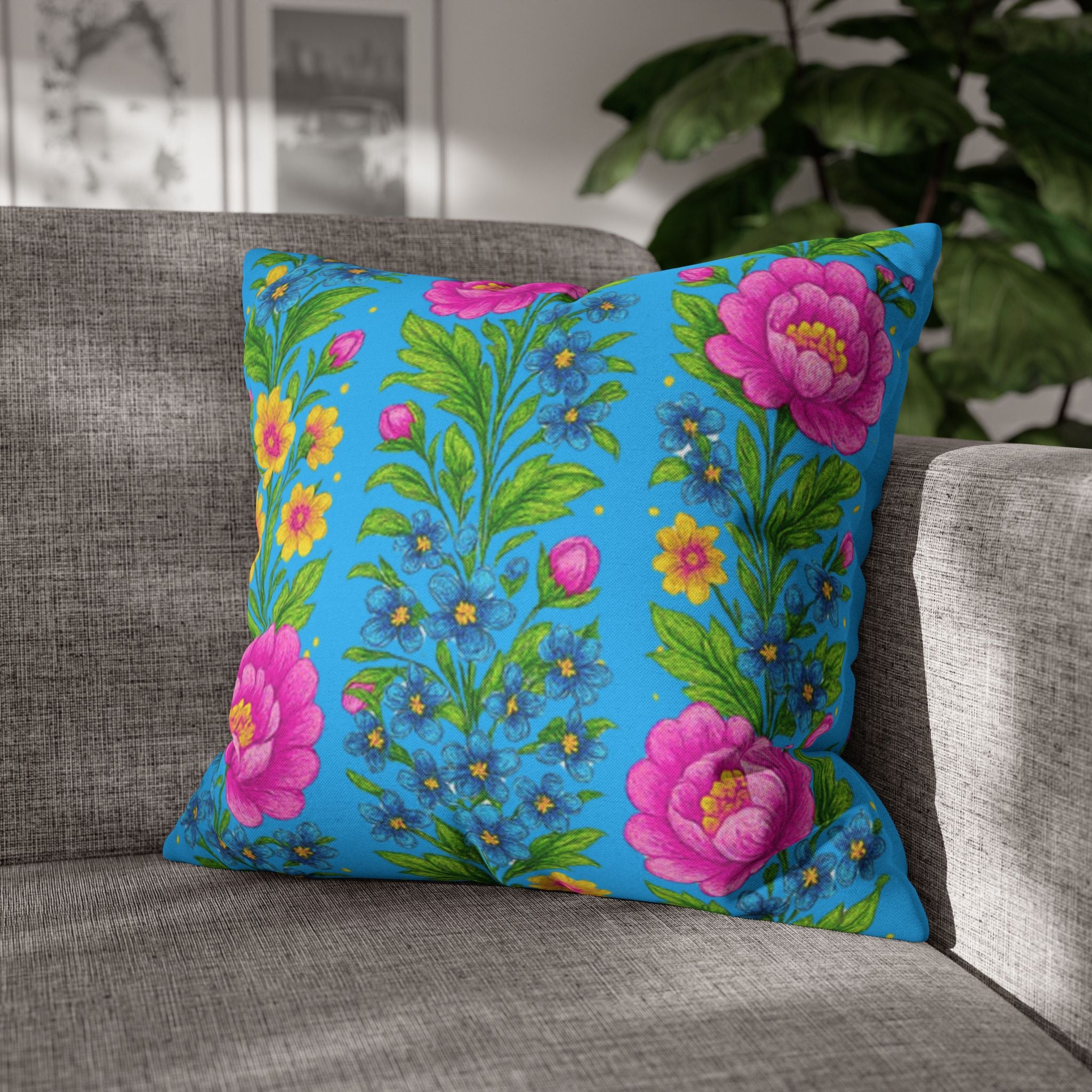 Square decorative pillowcase featuring vertical rows of pink peonies and yellow wildflowers on a bright sky-blue background, resting on a grey fabric sofa.