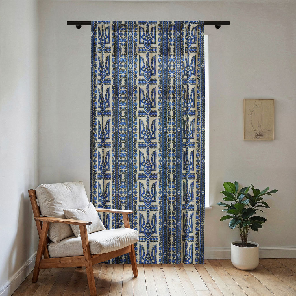 A light-filtering sheer window curtain featuring a vertical blue and black Ukrainian Trident (Tryzub) and folk embroidery pattern. The curtain is hanging in a bright, modern room next to a wooden armchair with cream cushions and a potted plant on a light wood floor.
