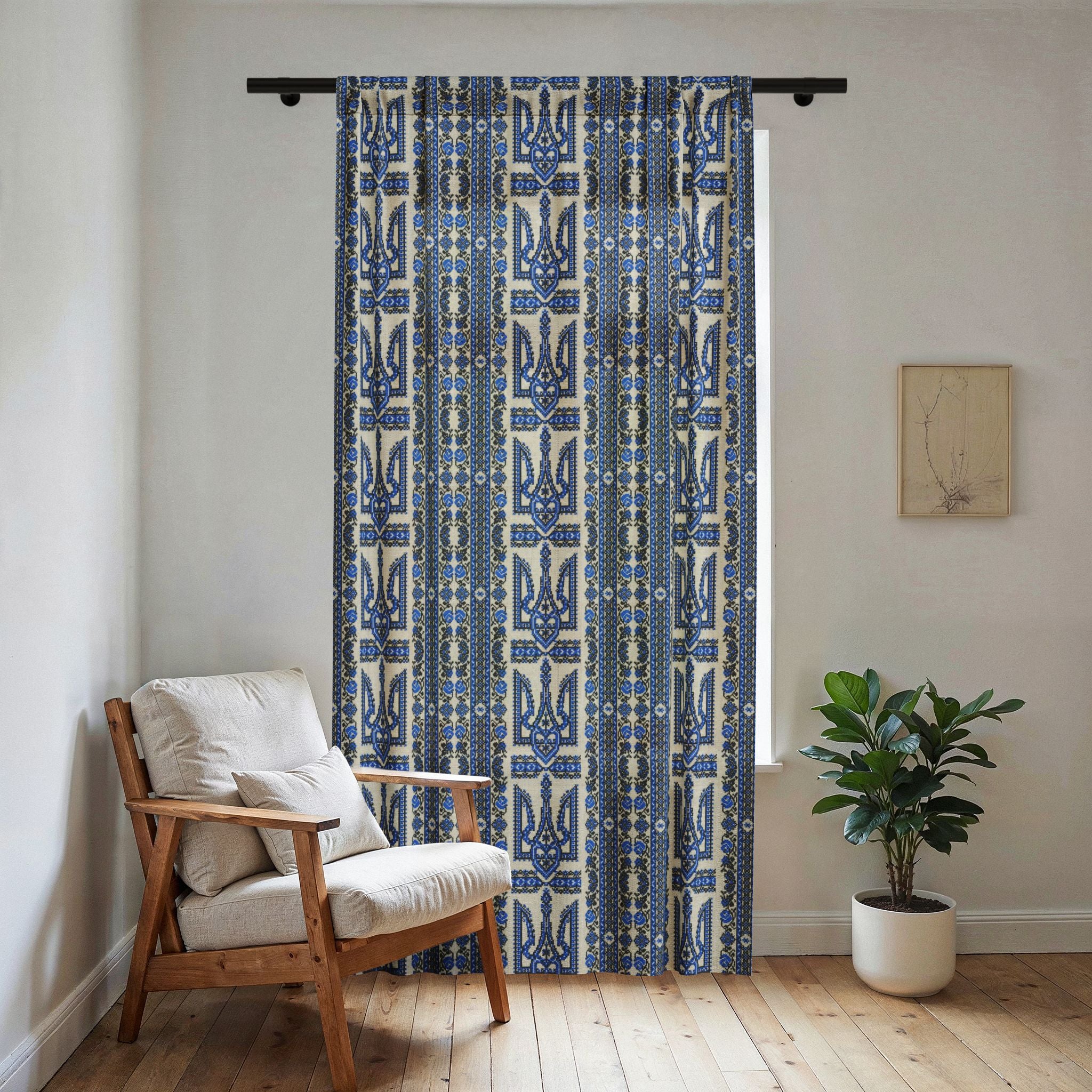 A light-filtering sheer window curtain featuring a vertical blue and black Ukrainian Trident (Tryzub) and folk embroidery pattern. The curtain is hanging in a bright, modern room next to a wooden armchair with cream cushions and a potted plant on a light wood floor.