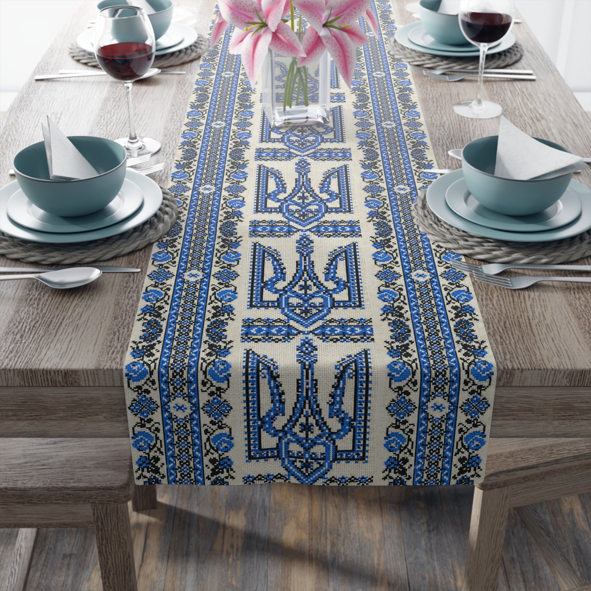 A high-angle view of a wooden dining table featuring a cream-colored table runner with a repeating blue and black Ukrainian Trident (Tryzub) and floral embroidery pattern. The table is set with blue bowls, wine glasses, and a vase of pink lilies.