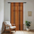 Vertical sheer window curtain featuring rust-brown and orange Trypillian symbols and stripes, filtering light next to a wooden armchair and potted plant.