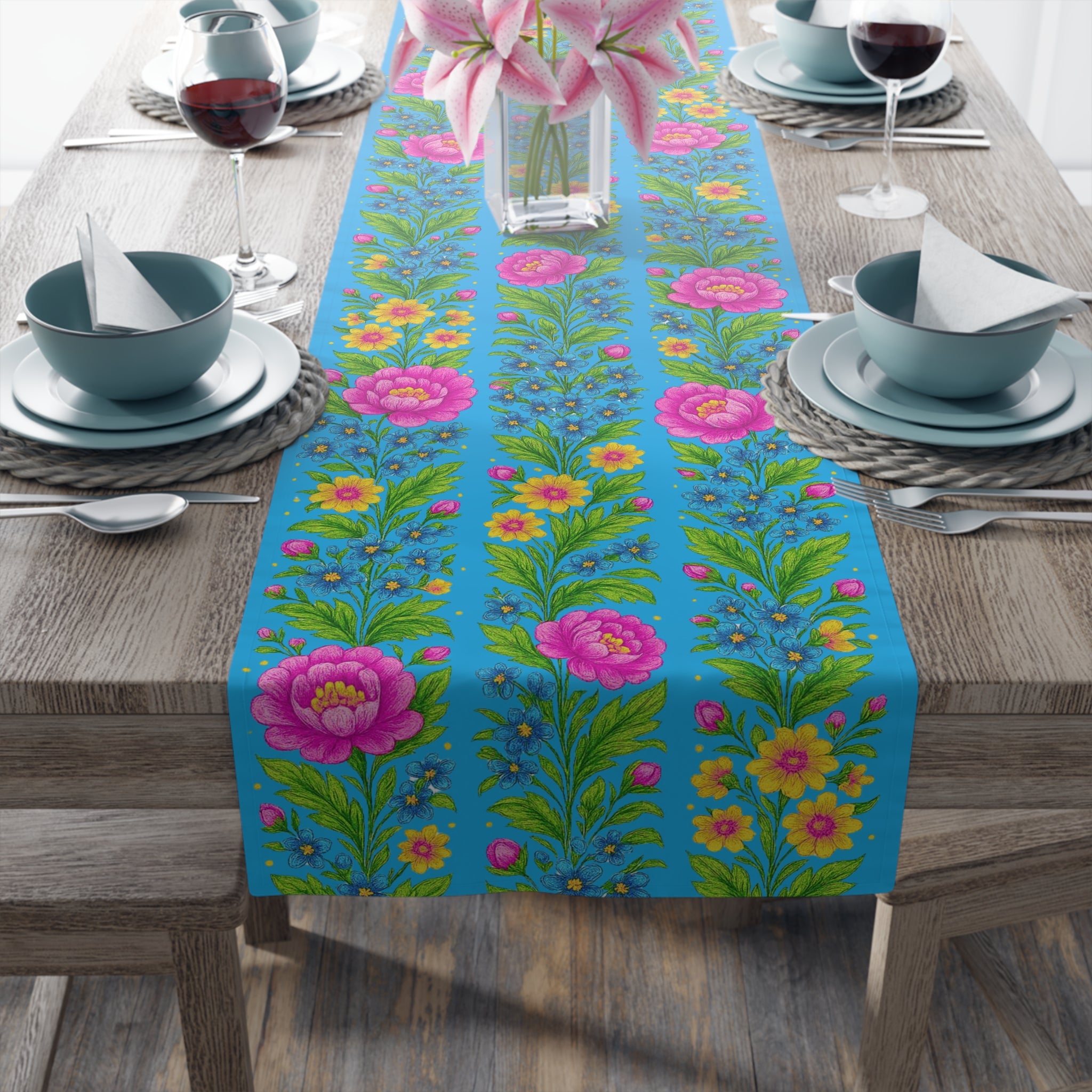 A bright sky-blue table runner with a repeating floral pattern of pink peonies and yellow wildflowers, displayed on a wooden dining table with elegant place settings.