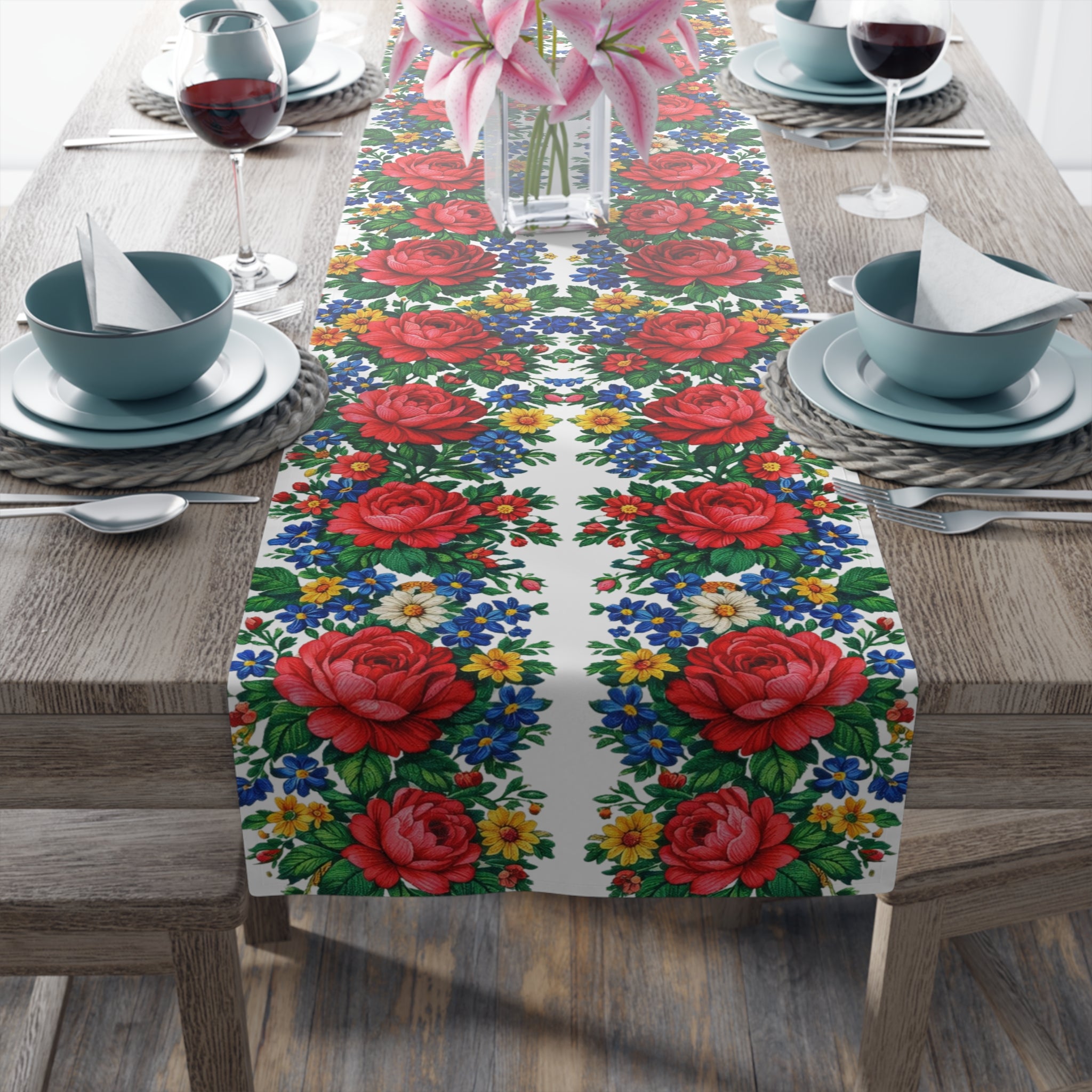 White table runner featuring a repeating border of pink roses and blue wildflowers, displayed on a wooden dining table with blue plates and wine glasses.