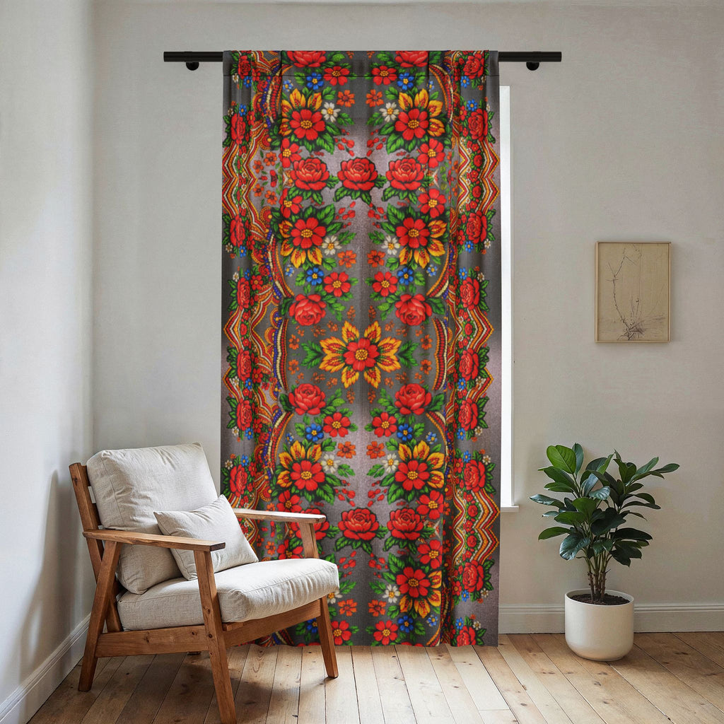 Sunlit room featuring the Mother’s Bloom sheer window curtain with a vibrant red and orange Ukrainian folk pattern on a soft grey background, hanging next to a wooden armchair and a white wall.