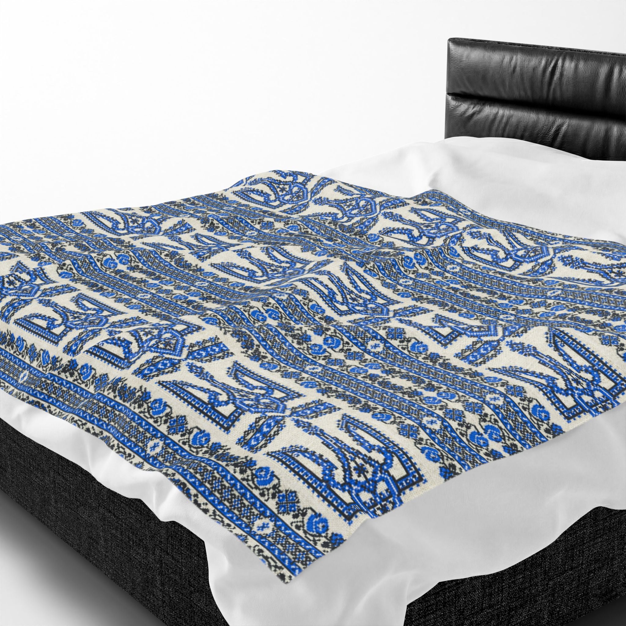 A soft velveteen plush blanket featuring a dense, repeating blue and black Ukrainian Trident (Tryzub) and floral embroidery-style pattern on a white background. The blanket is draped over a modern black bed frame with white sheets.
