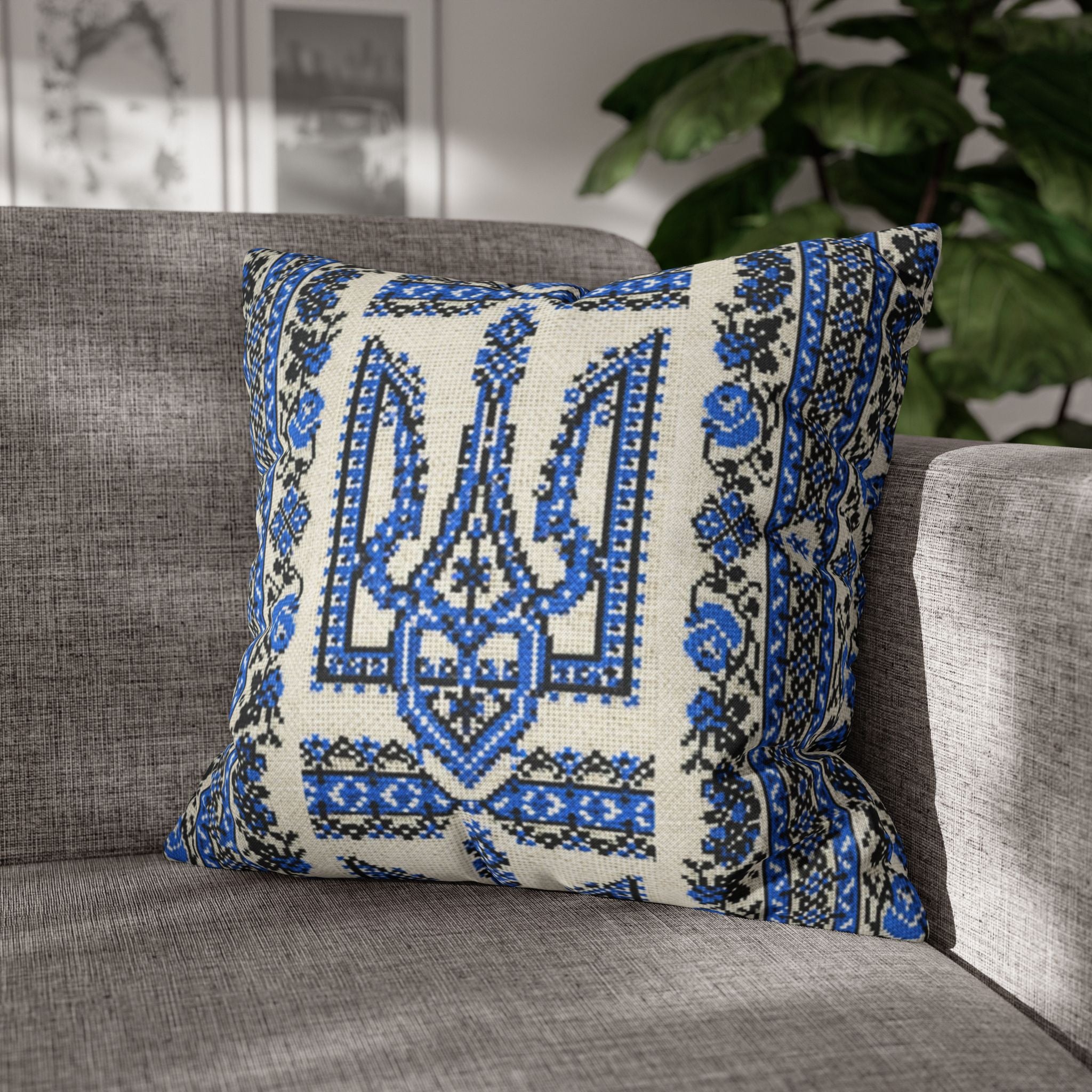 A square decorative pillow sitting on a grey fabric sofa. The pillow features a cream background with a large, intricate blue and black printed Ukrainian Trident (Tryzub) in the center, surrounded by traditional folk-style floral borders and patterns.