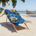 Midnight Symphony beach towel with large blue and cream floral patterns draped over a wooden lounge chair on a tropical sandy beach with palm trees and a boat in the background.