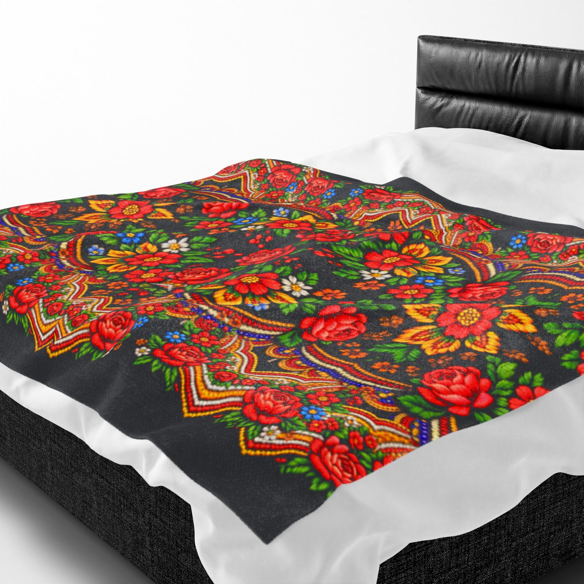 A plush velveteen blanket named "Mother’s Bloom" with a vibrant red and yellow Ukrainian folk floral pattern, draped over a white bed with a black leather headboard.