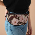 A person wearing the Sakura Noir fanny pack with a pink cherry blossom and dark brown leaf pattern over blue jeans and a black t-shirt.