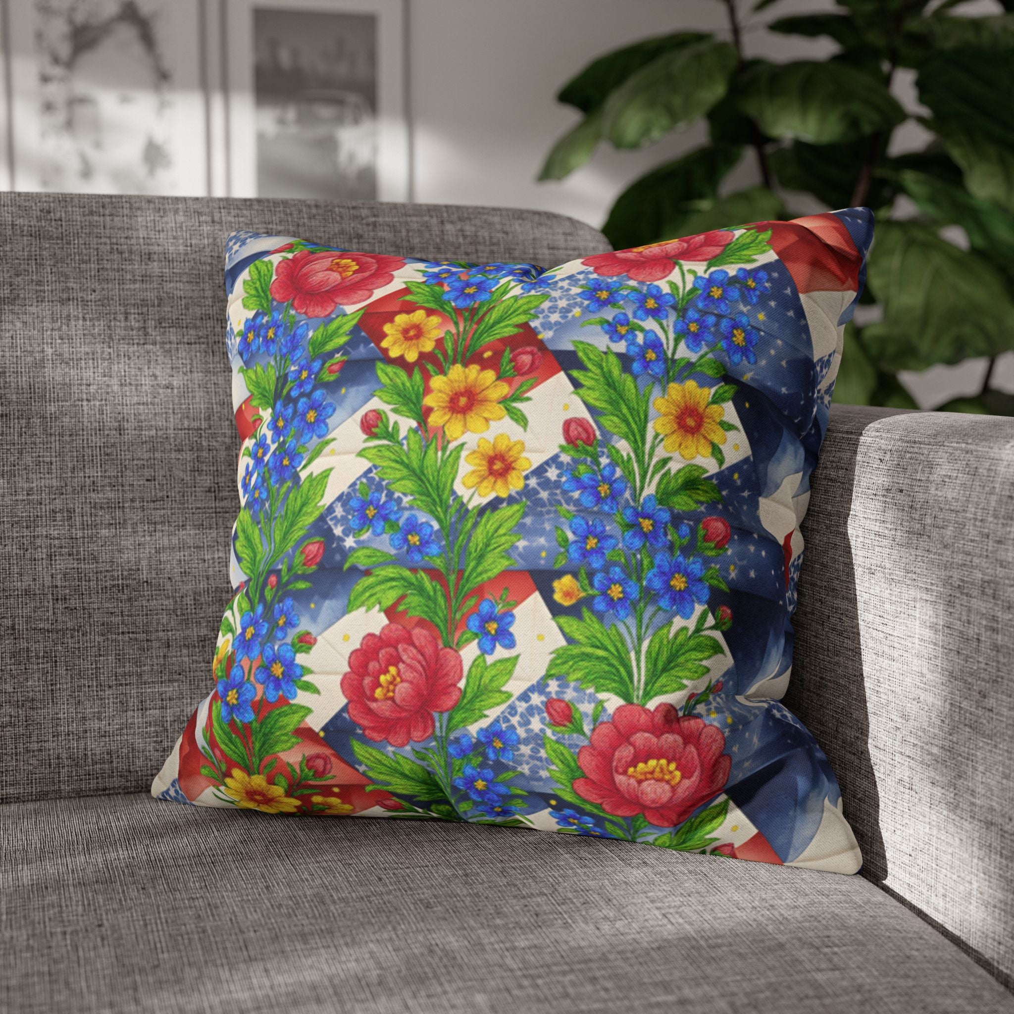 Square decorative pillow titled American Cozy, featuring red roses and blue wildflowers over a blue geometric pattern, resting on a grey fabric sofa.