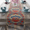 A high-quality table runner with a vintage Americana design. It features repeating red and blue floral medallions and intricate folk patterns on a cream-colored background, laid across a rustic wooden dining table.